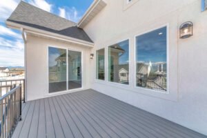 Deck of a house with reflective sliding glass door and picture windows. Home exterior with wall lights, wooden planks flooring and black metal railings.